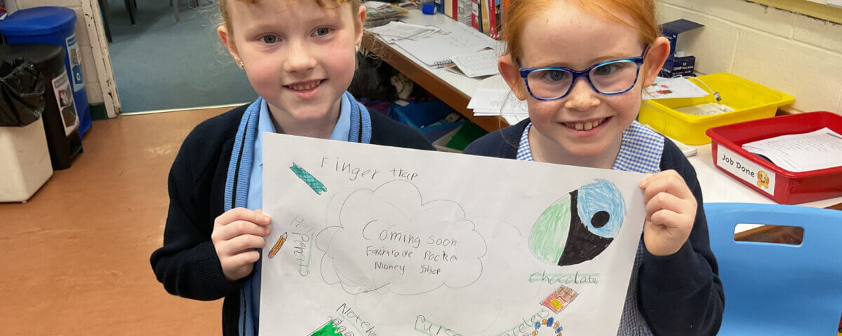 Two young girls in school uniforms smile and hold up a colorful drawing titled "Finger tap," which features doodles of chocolate, music notes, a lock, and other objects. They are in a classroom setting.