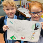 Two young girls in school uniforms smile and hold up a colorful drawing titled "Finger tap," which features doodles of chocolate, music notes, a lock, and other objects. They are in a classroom setting.