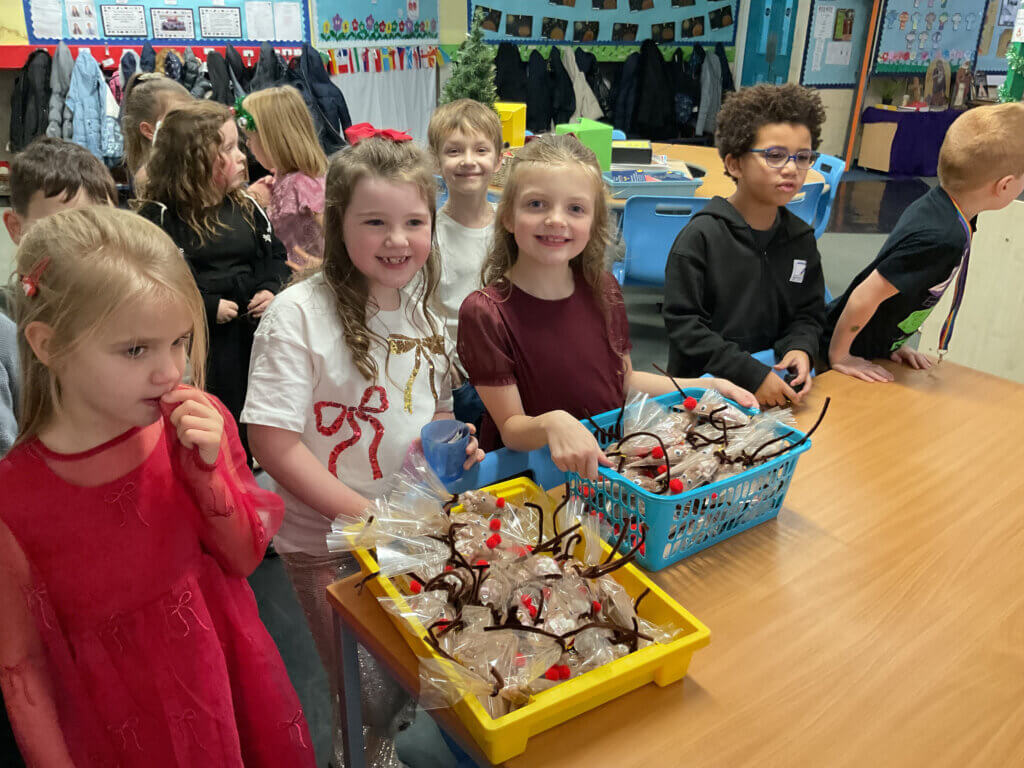 A group of young children in festive outfits stand around a table with trays of reindeer-shaped treats in a decorated classroom, smiling and appearing excited.
