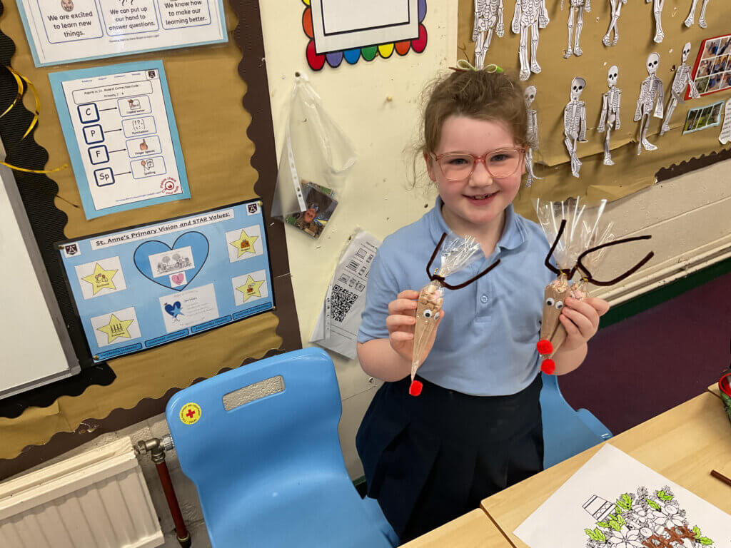 A young girl with glasses smiles while holding two reindeer-themed treats made from clear bags, brown pipe cleaners, googly eyes, and red pom-poms. She is sitting in a classroom with colorful decorations and drawings.