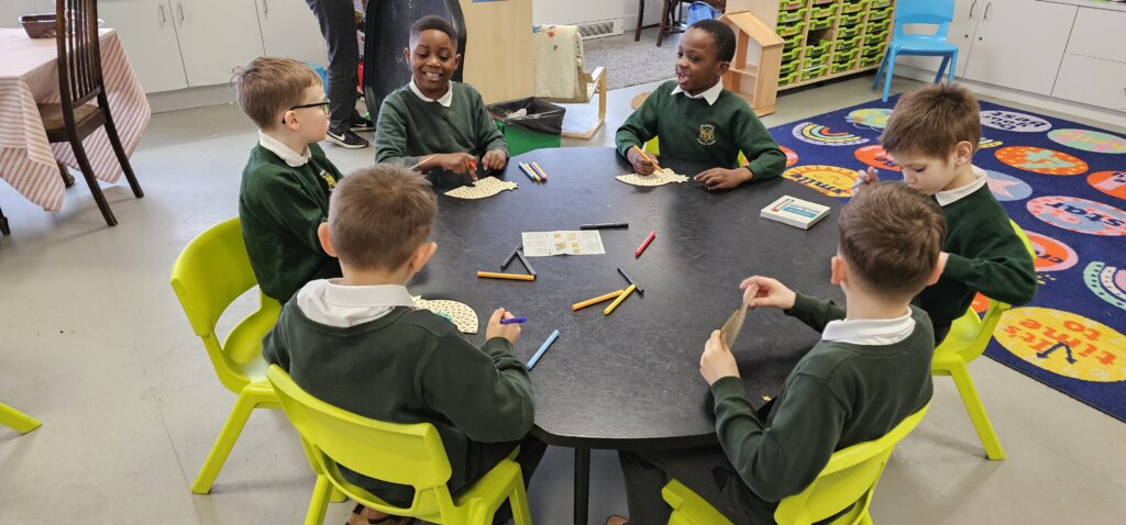 Six young boys in green school uniforms sit around a black table in a classroom, coloring on circular pieces of paper with markers. The room has colorful chairs and a bright rug with playful designs.