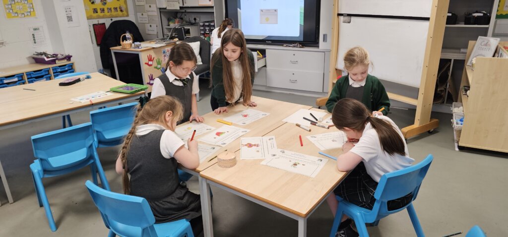 Five young girls sit around a classroom table, drawing and coloring on worksheets with markers. The classroom has bright chairs, wooden tables, and educational materials visible in the background.