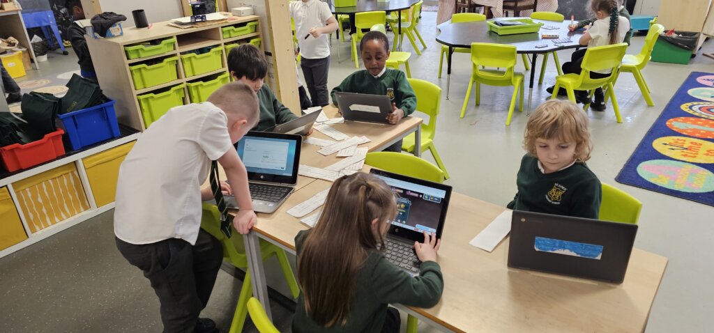 A group of young children in a classroom sits and stands around tables, focused on using laptops. Bright green chairs and classroom materials are visible in the background.