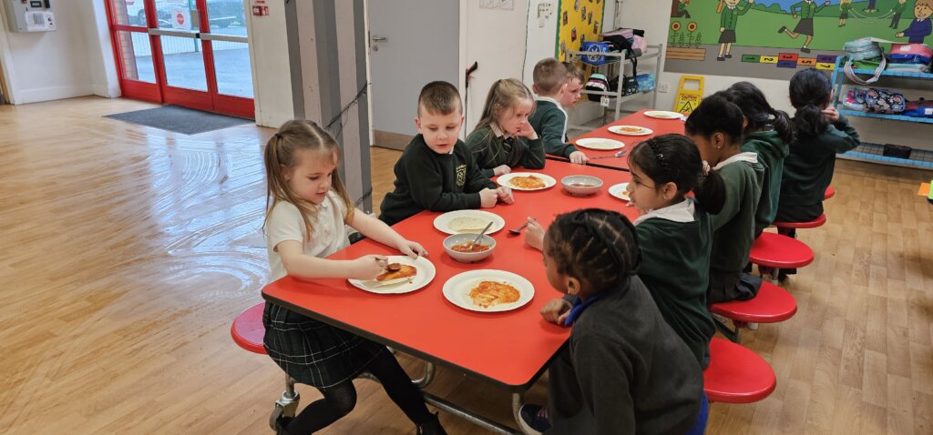 Young children in school uniforms sit around a red table in a classroom, making or eating food on plates. Ingredients and utensils are on the table, and colorful wall decorations are visible in the background.