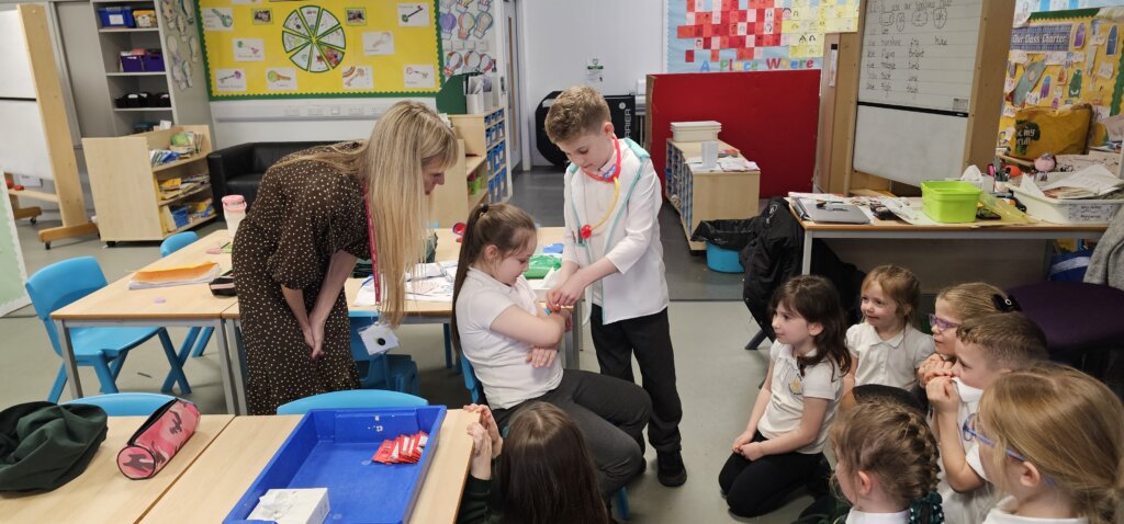 A group of young children sit on the classroom floor watching as a boy pretends to use a toy stethoscope on a seated girl. An adult woman stands nearby, observing and smiling. The classroom is colorful and lively.