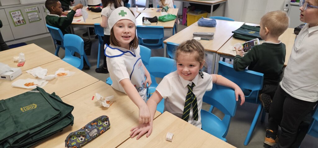 Two young girls in a classroom, one dressed as a nurse placing a bandage on the other's hand. Other children sit at desks in the background. The setting is bright and cheerful.