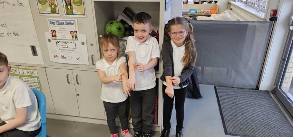 Three young children stand indoors, smiling and showing their arms, as if displaying something on their skin. They are dressed in school uniforms, standing in front of a cabinet with toys and posters. Another child sits to the left.