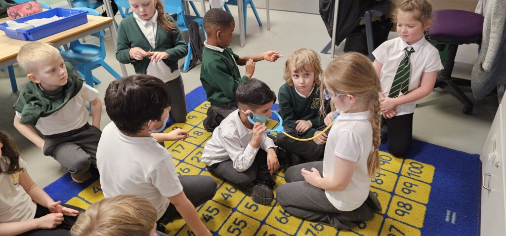 A group of young children in school uniforms sit and kneel on a yellow classroom mat numbered 1 to 100, taking turns listening to a stethoscope during a class activity.