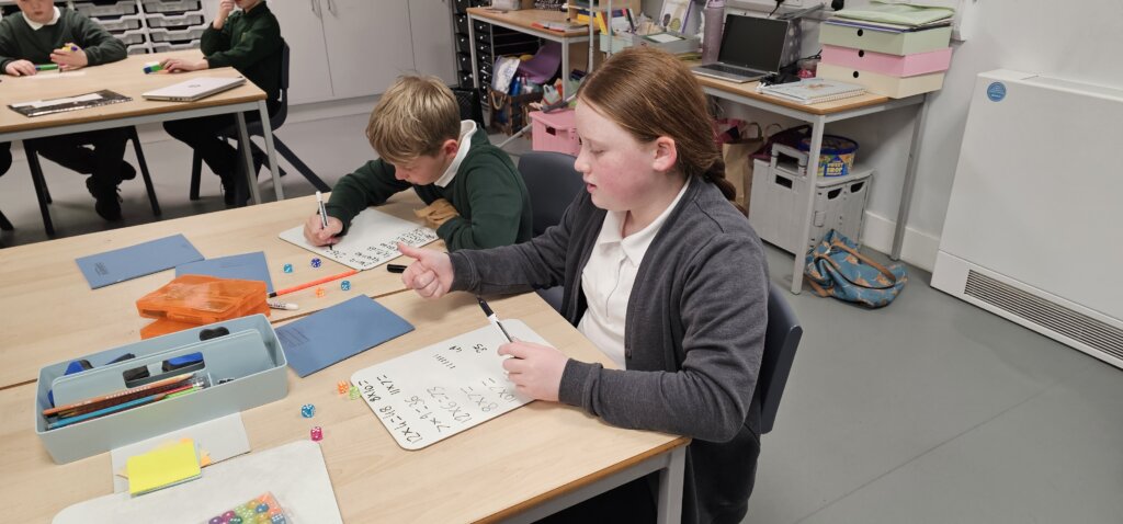 Two children sit at a classroom table, writing on whiteboards with markers. Various stationery, folders, and dice are scattered on the table. Other students work at desks in the background.