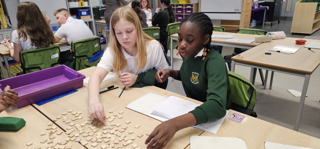 Two girls sit at a classroom table, sorting wooden letter tiles. One girl wears a white shirt and reaches for a tile, while the other, in a green sweater, talks. Other students work at desks in the background.