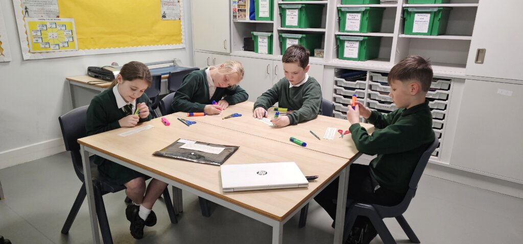 Four children in green school uniforms sit around a classroom table, focused on writing or drawing with colorful markers. The room has shelves with green bins, and a laptop and supplies are on the table.