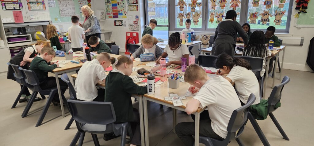 A classroom with students sitting at tables, working on assignments. Two adults assist them. The walls are decorated with colorful artwork and posters, and large windows let in natural light.