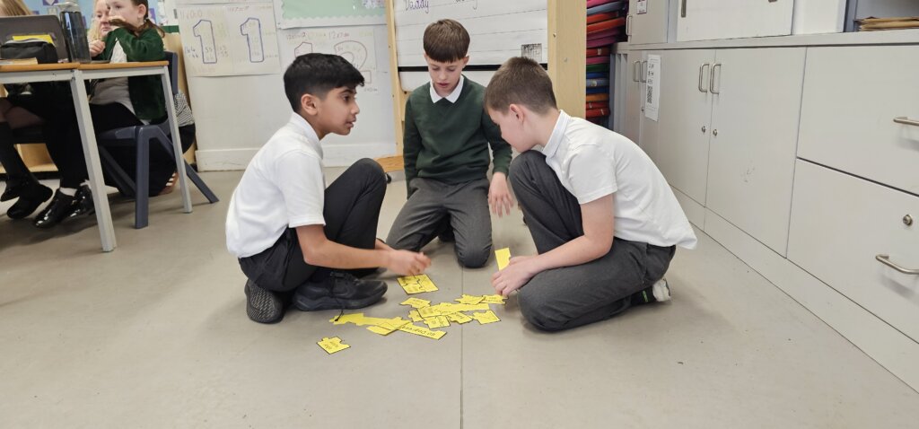 Three boys sit on the classroom floor, working together to arrange yellow cards. Other students and classroom furniture are visible in the background.