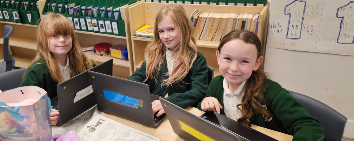 Three young girls in school uniforms sit at a classroom table, smiling at the camera. Each has a laptop in front of her, and the table is scattered with papers, pencils, and a plush toy. Classroom shelves and charts are visible behind them.