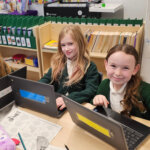 Three young girls in school uniforms sit at a classroom table, smiling at the camera. Each has a laptop in front of her, and the table is scattered with papers, pencils, and a plush toy. Classroom shelves and charts are visible behind them.