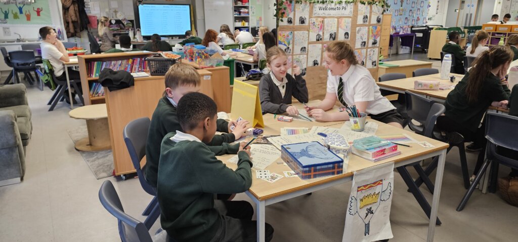 A group of schoolchildren in uniform sit around tables working on arts and crafts projects in a colorful, busy classroom filled with books, drawings, and educational materials.