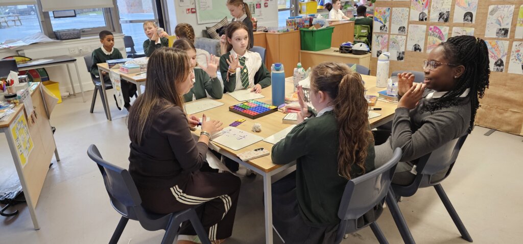 A group of schoolchildren and a teacher sit around a classroom table, playing a colorful board game. Other students are visible working and talking in the background. The classroom walls display various student artwork.