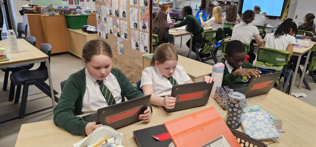 Three schoolchildren in uniform are sitting at a classroom table, focused on using tablets. Other students and a teacher are in the background, with colorful artwork displayed on the wall and various school supplies on the table.