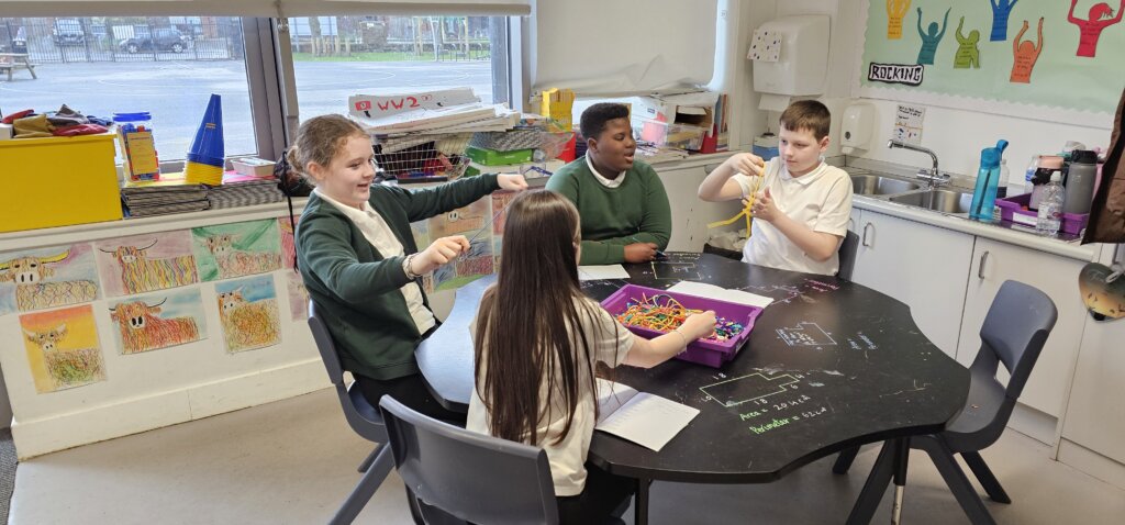Four children sit around a black table in a classroom, playing with colorful string or pipe cleaners. Bright animal artwork decorates the wall, and shelves with supplies are visible in the background. The children are smiling and engaged.