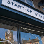 A sign reading "START-UP STREET" above a building entrance, with the reflection of a clock tower and blue sky visible in the glass door below.