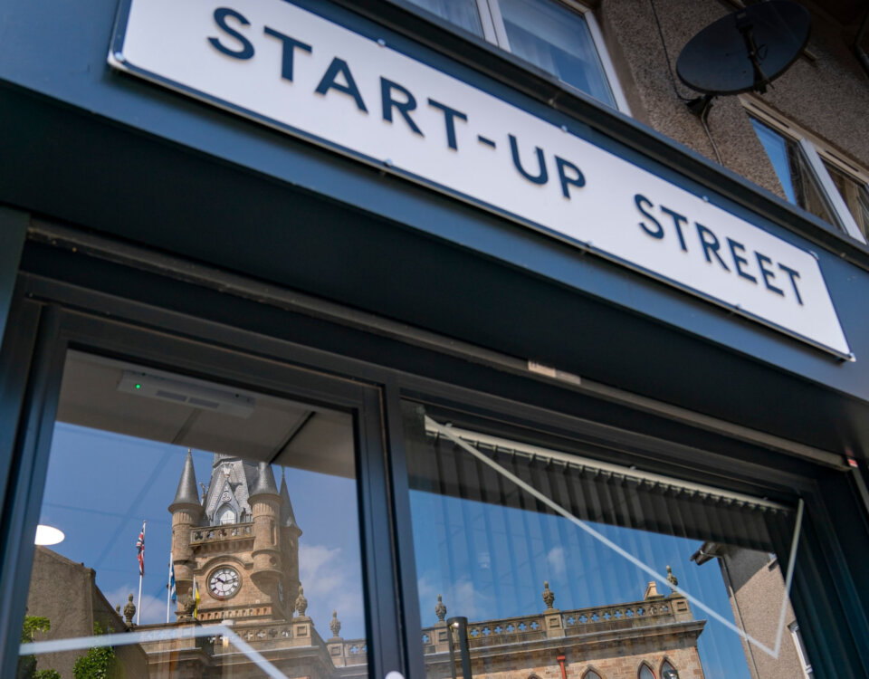 A sign reading "START-UP STREET" above a building entrance, with the reflection of a clock tower and blue sky visible in the glass door below.