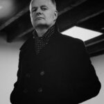 A person with short, swept-back hair wearing a dark double-breasted coat and a polka dot scarf looks confidently at the camera; exposed wooden ceiling beams are visible above. The image is in black and white.