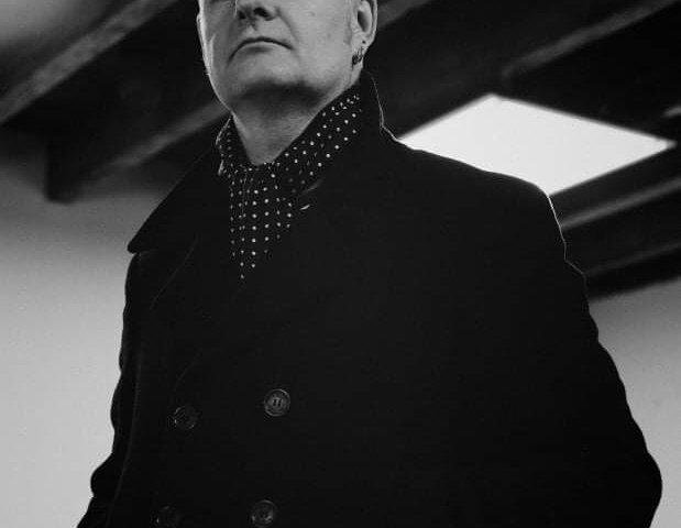 A person with short, swept-back hair wearing a dark double-breasted coat and a polka dot scarf looks confidently at the camera; exposed wooden ceiling beams are visible above. The image is in black and white.