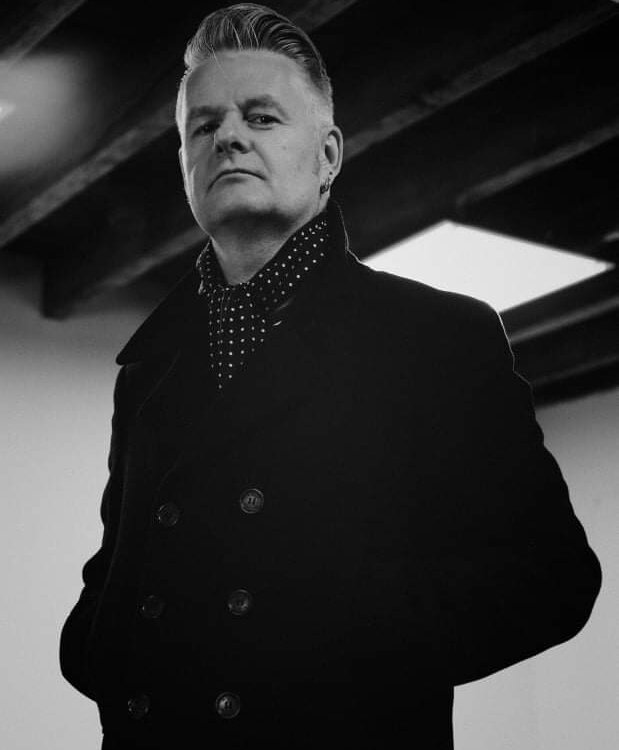 A person with short, swept-back hair wearing a dark double-breasted coat and a polka dot scarf looks confidently at the camera; exposed wooden ceiling beams are visible above. The image is in black and white.