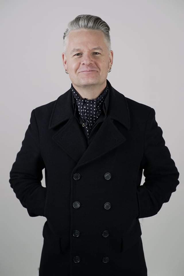 A person with short, gray, slicked-back hair is wearing a black double-breasted coat and a black scarf with white polka dots, standing against a plain light background with hands behind their back.