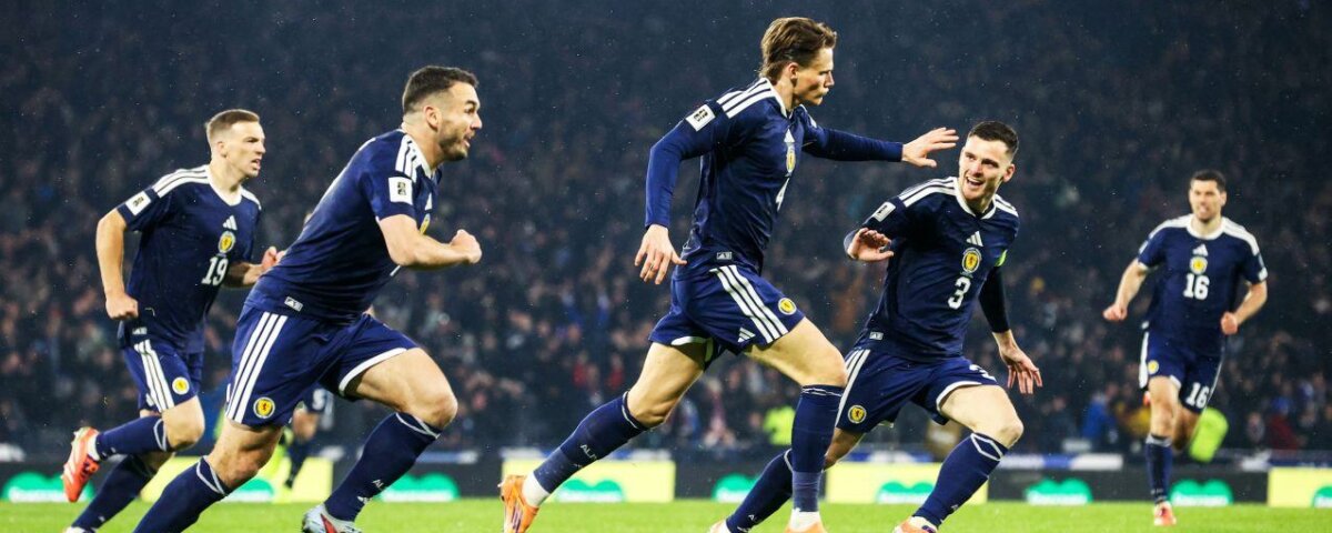 A group of soccer players in navy blue uniforms celebrate on the field, smiling and running with arms outstretched, with a crowd of spectators in the background.