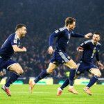 A group of soccer players in navy blue uniforms celebrate on the field, smiling and running with arms outstretched, with a crowd of spectators in the background.
